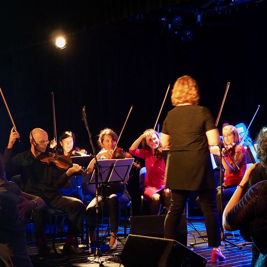 Anna Kalska dyryguje orkiestrą Formacji Tango Para Rodos. Na zdjęciu widać skrzypków kończących frazę muzyczną z podniesionymi smyczkami.