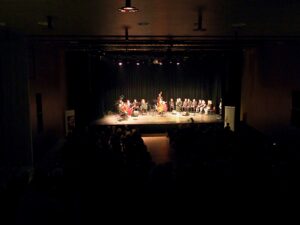Ciemna widownia sali koncertowej, w oddali scena na której znajduje się chór i orkiestra Formacji Tango Para Rodos