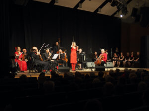 Na scenie Agnieszka Kowlskka przeprowadza z publicznością rozgrzewkę wokalną. W tle siedzi Formacja Tango Para Rodos