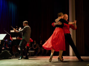 Chór i orkiestra Formacji Tango Para Todos pod dyrekcją Grzegorza Mańkowskiego. Na pierwszym planie tango tańczy para taneczna - tancerka w czerwonej powiewającej spódnicy, partner w czerwonej koszuli i czarnych spodniach.
