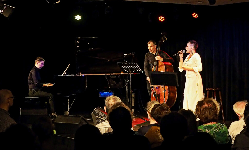 Na scenie Promu Kultury Saską Kępa trio Sugar Jazz w składzie: wokalistka ubrana na biało, pianista i kontrabasista ubrani na czarno.