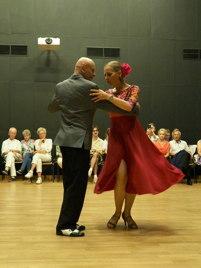 Tango tańczy Tomasz Sadlakowski ubrany w czarne spodnie i szarą marynarkę i Monika Kwiatkowska ubrana w czerwoną, rozkloszowaną sukienkę. Na głowie ma wpięty czerwony kwiat.