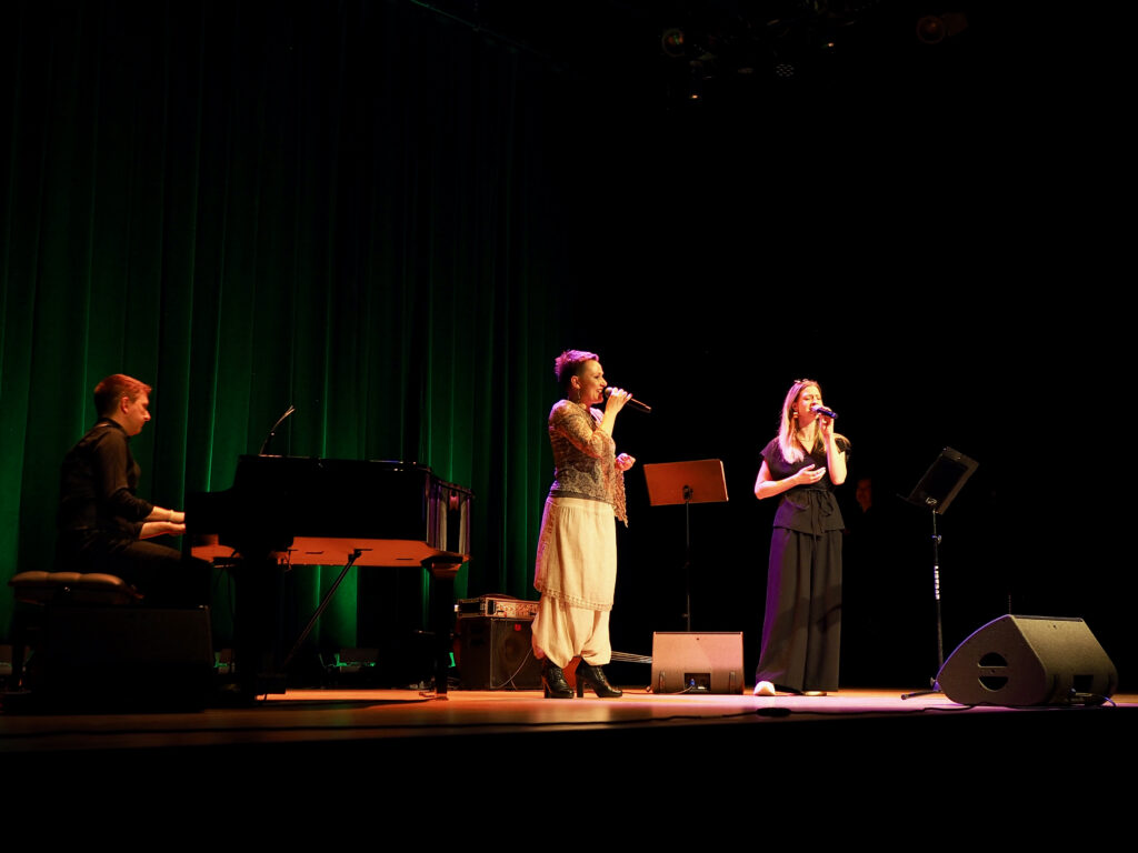 Na przyciemnionej scenie widać pianistę, Agnieszkę Kowlaska - wokalistkę i zaproszoną koleżankę