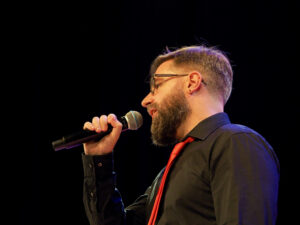 Śpiewa Duarte Peralta. Jest ubrany w czarną koszulę i czerwony krawat