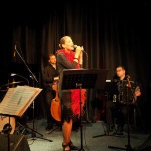 Na scenie śpiewa wokalistka, w tle kontrabasista i akordeonista